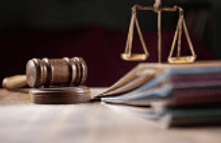 A judge's gavel resting on a wooden block next to an open book with visible pages. In the background, a set of balanced scales symbolizes justice.